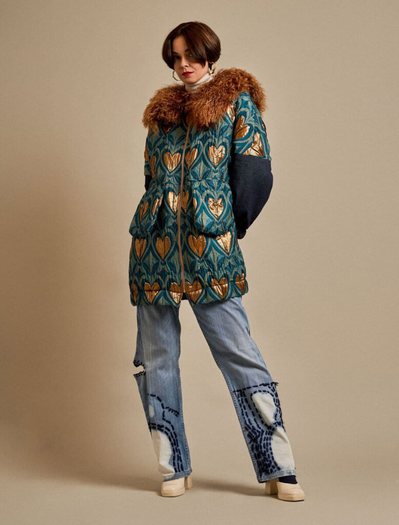 Mujer luciendo el Abrigo Raxao con cuello de piel natural y estampado de corazones dorados sobre fondo azul.